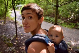 Woman hiking in the woods with a baby strapped to her back.
